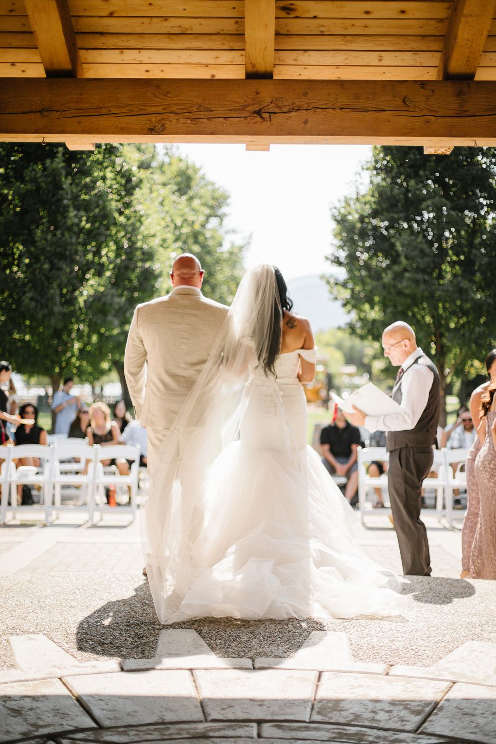 Wedding guests seated under a wooden pavilion during an outdoor ceremony.