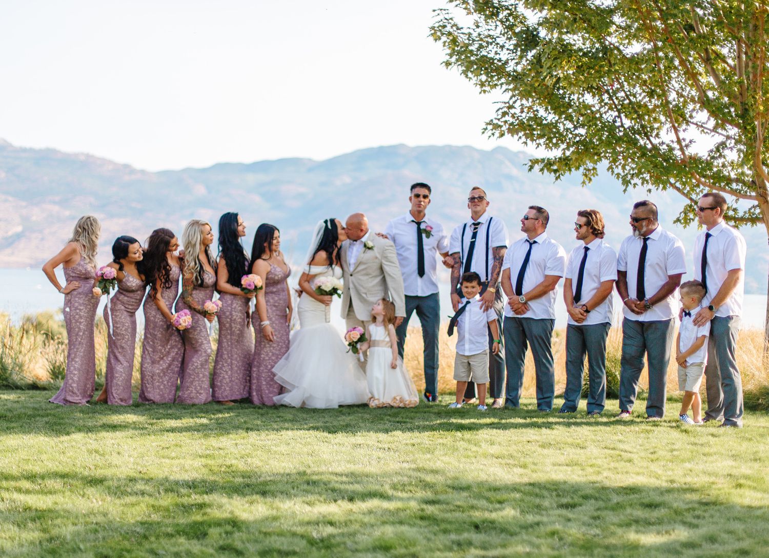 Large wedding party gathered on a grassy hillside for formal group photos.