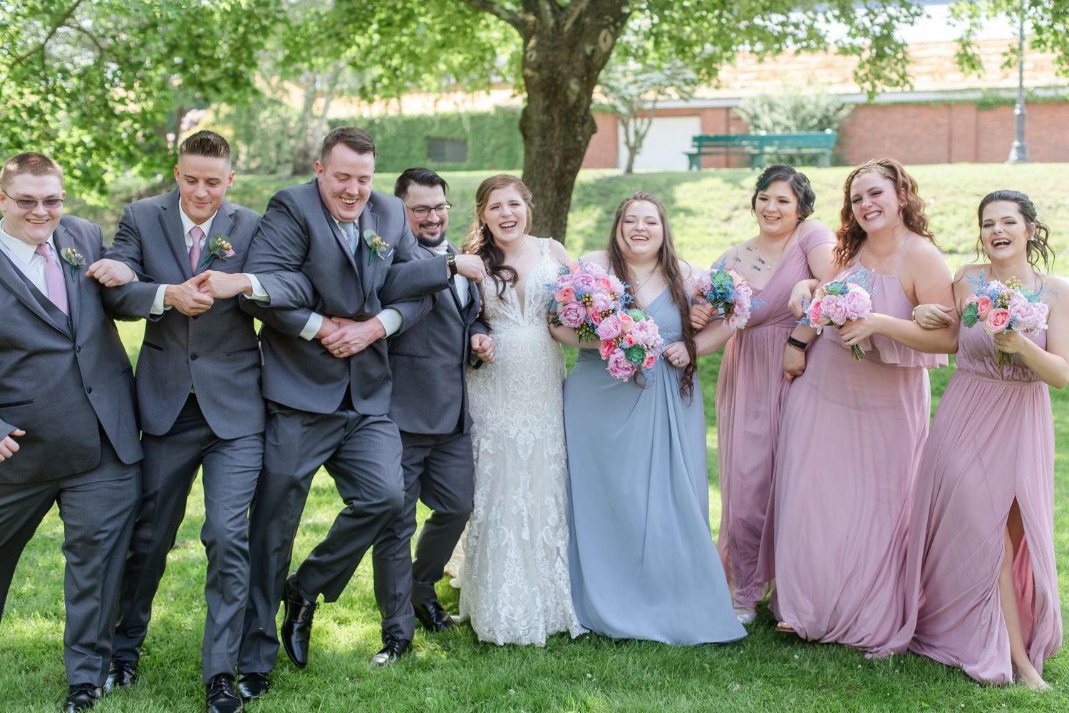 Wedding party members gather for group photos in gray and mauve attire under lush trees.