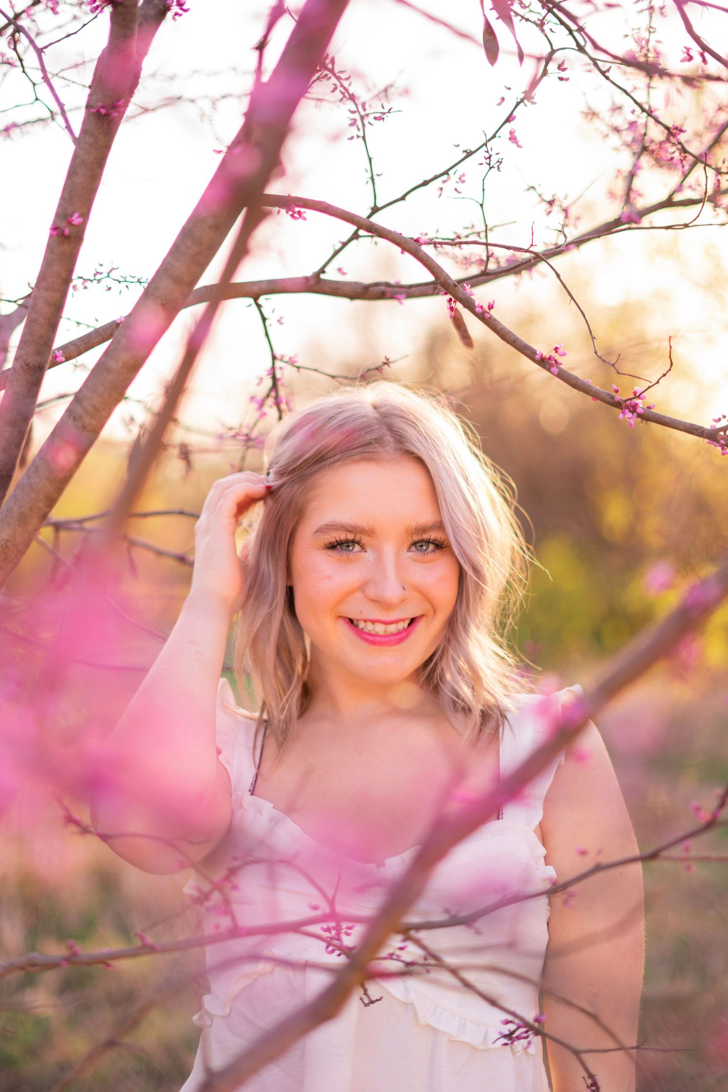 Blonde woman smiling among pink blooming tree branches in warm golden hour light.