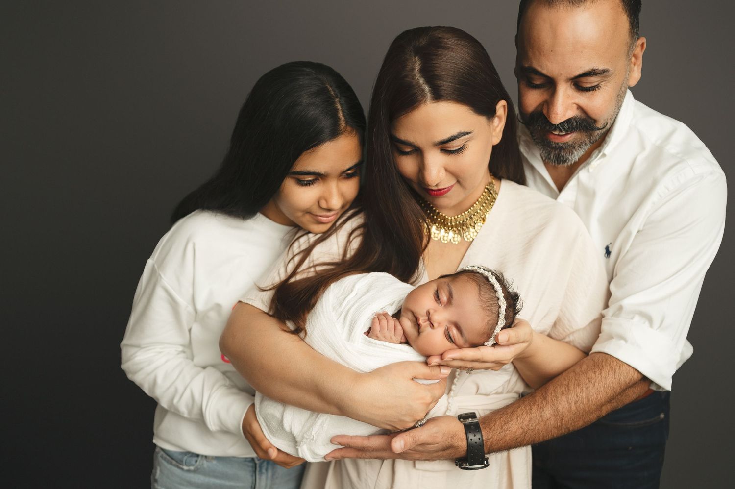 Tender family portrait showing affectionate parents and older child cuddling with sleeping newborn against dark background.
