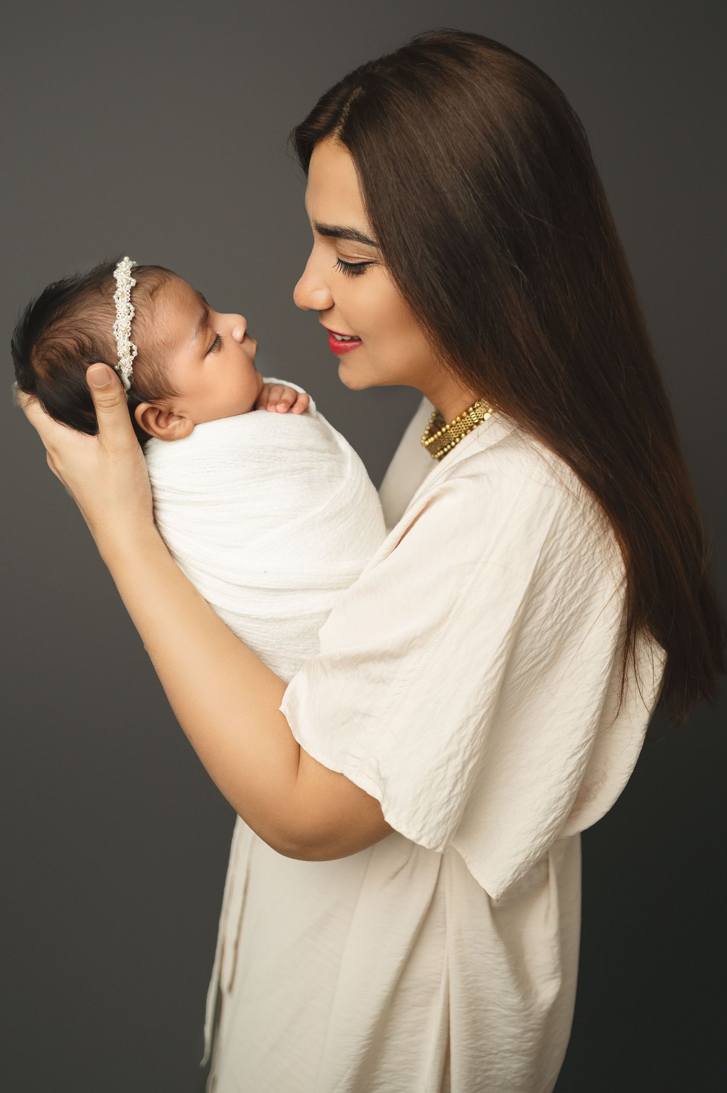 Woman in white wearing gold jewelry holds sleeping newborn wrapped in white against dark background.