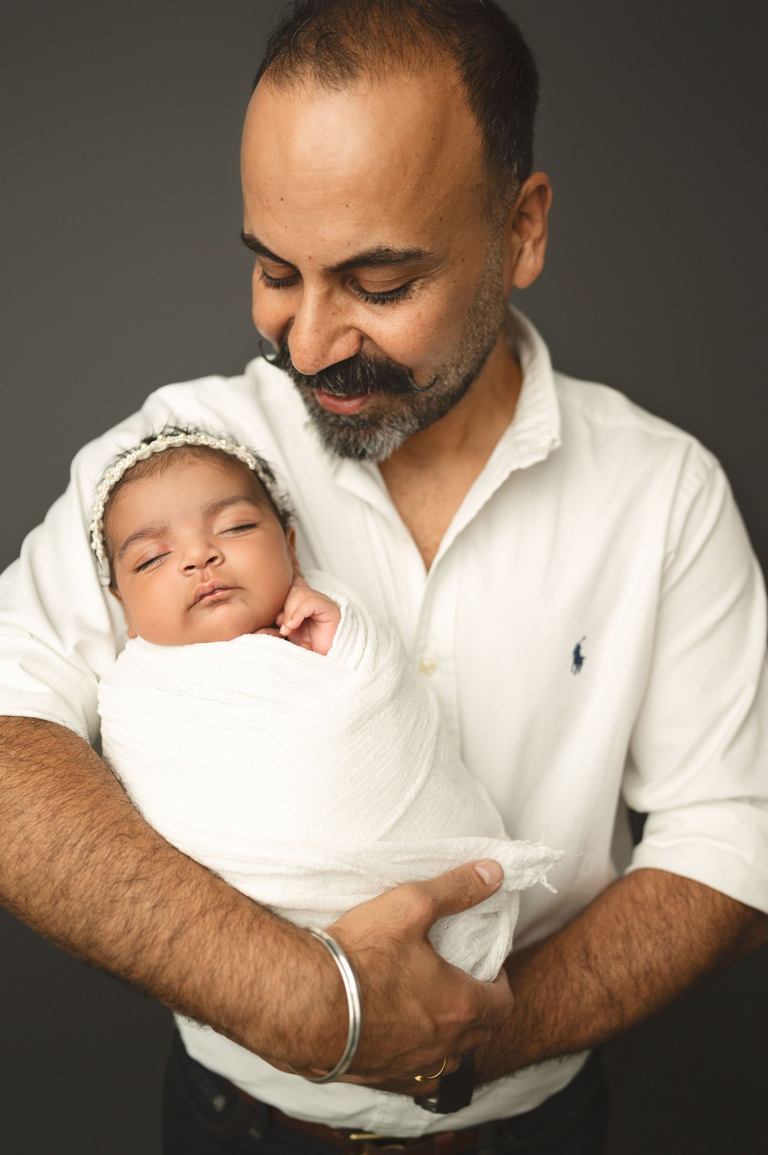 Bearded parent in white shirt smiles lovingly while holding peaceful sleeping newborn swaddled in white.
