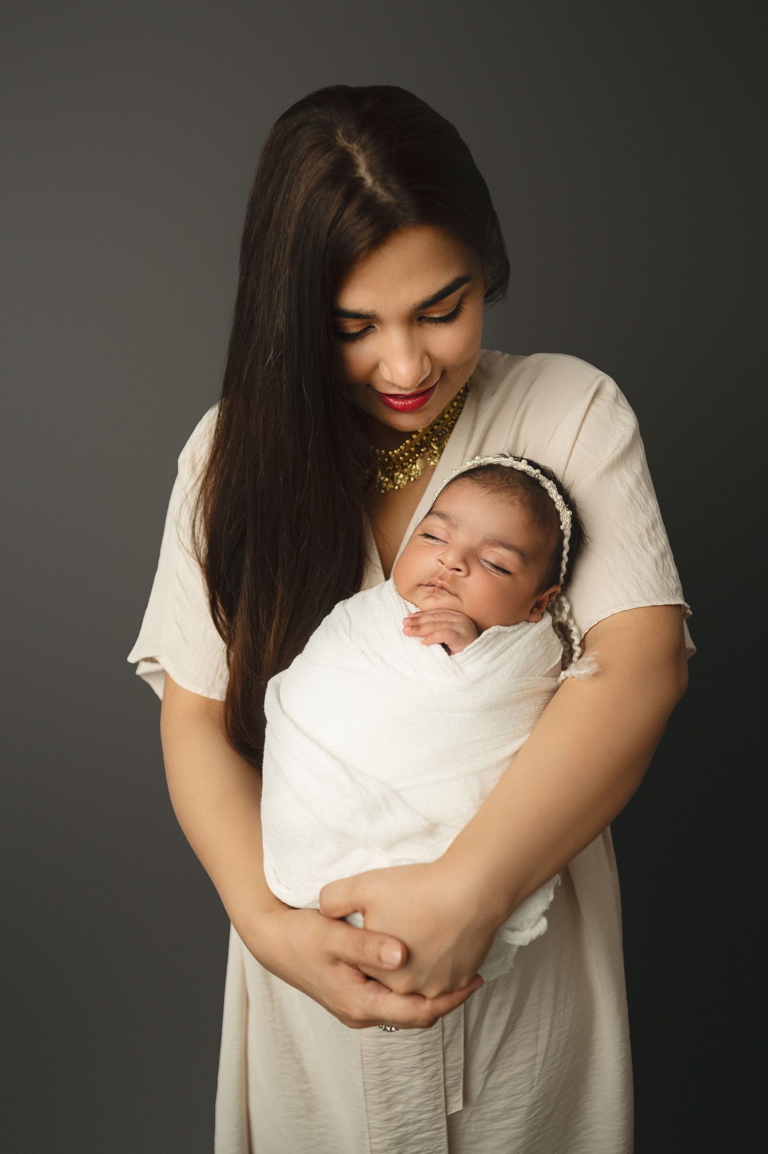 Woman in light dress cradles sleeping newborn wearing delicate headband against dark backdrop.