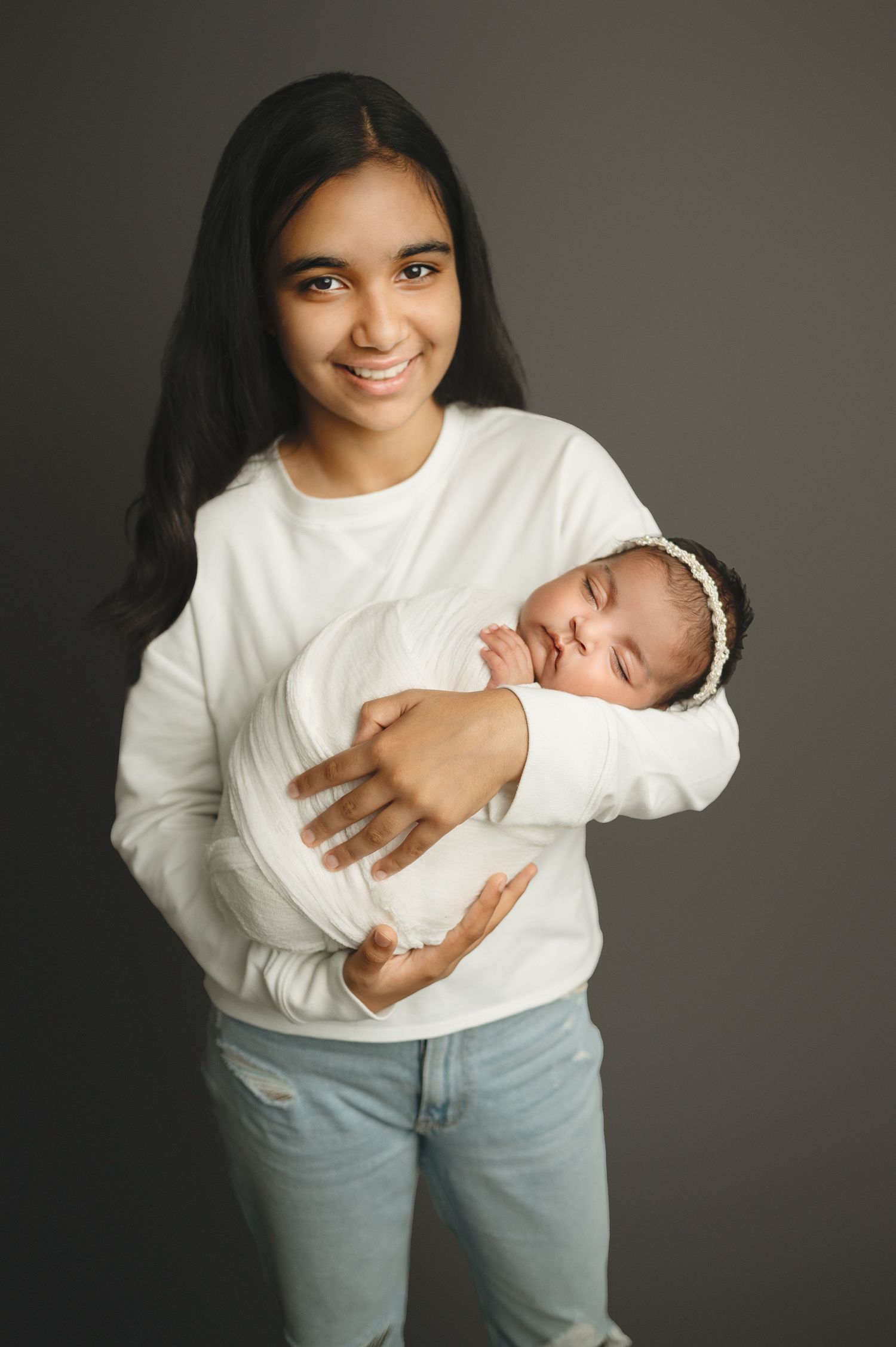 Young woman in casual white top and jeans holds sleeping swaddled newborn against grey backdrop.