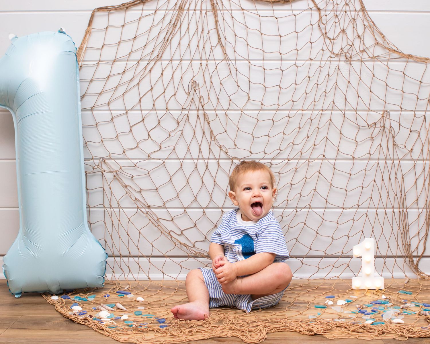 Baby boy sits on fishing net beside a blue number 1 balloon celebrating his first birthday party.