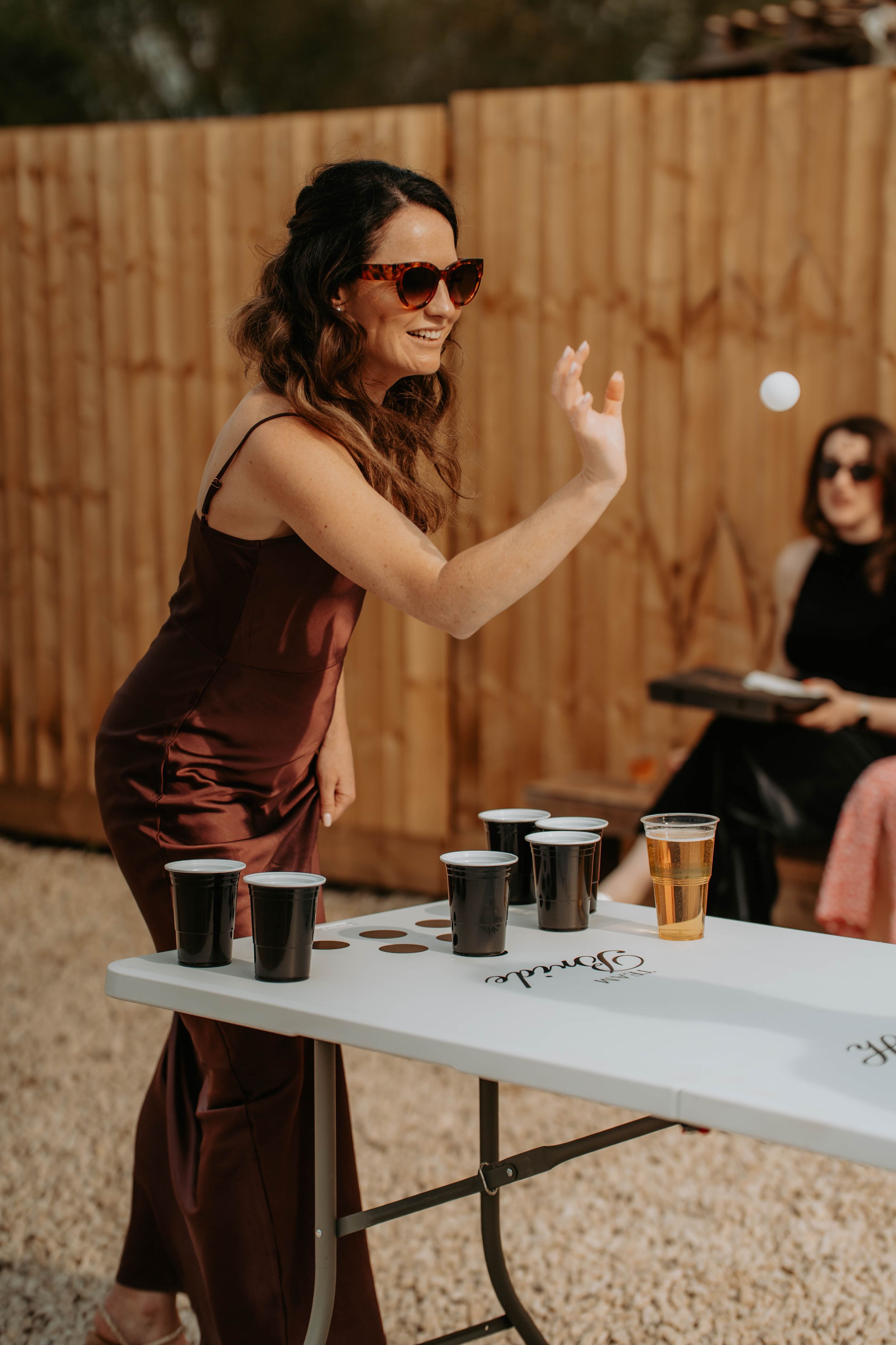 Woman in burgundy dress playing beer pong at outdoor bachelorette party, tossing ping pong ball toward cups.