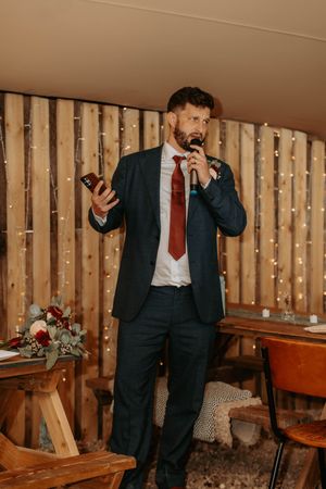 Bearded groom in navy suit and red tie gives speech holding microphone and phone at rustic wedding venue.