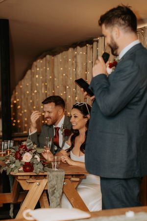 A bride and groom laugh joyfully at their wedding reception while a groomsman delivers a heartfelt speech.