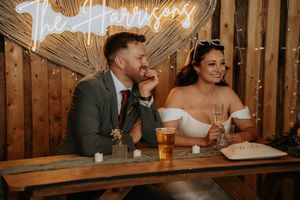 Bride and groom laughing at sweetheart table with neon Harrison sign and fairy lights at rustic wedding reception.