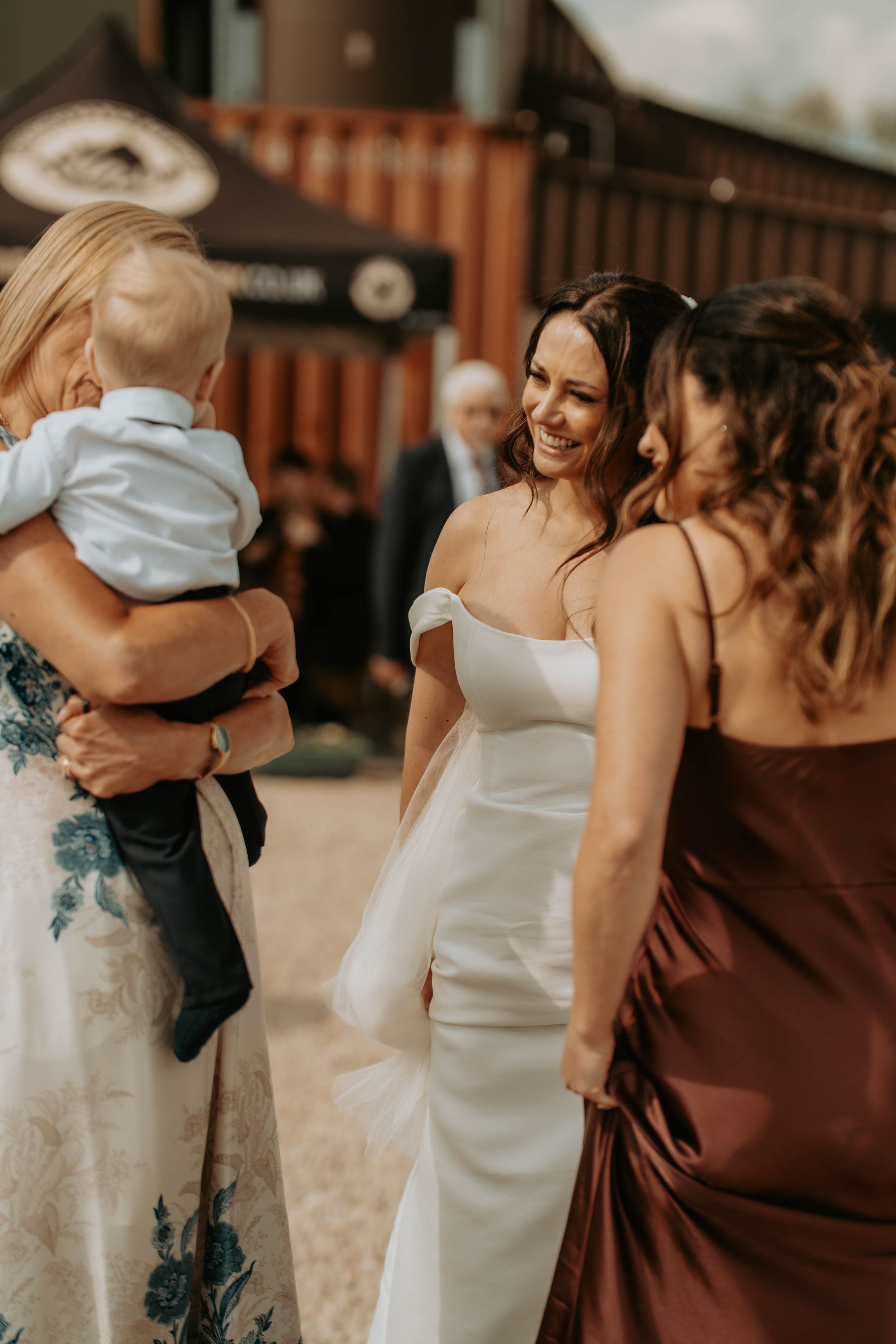 Bride in white off-shoulder gown laughing with bridesmaid in burgundy dress at outdoor wedding venue.