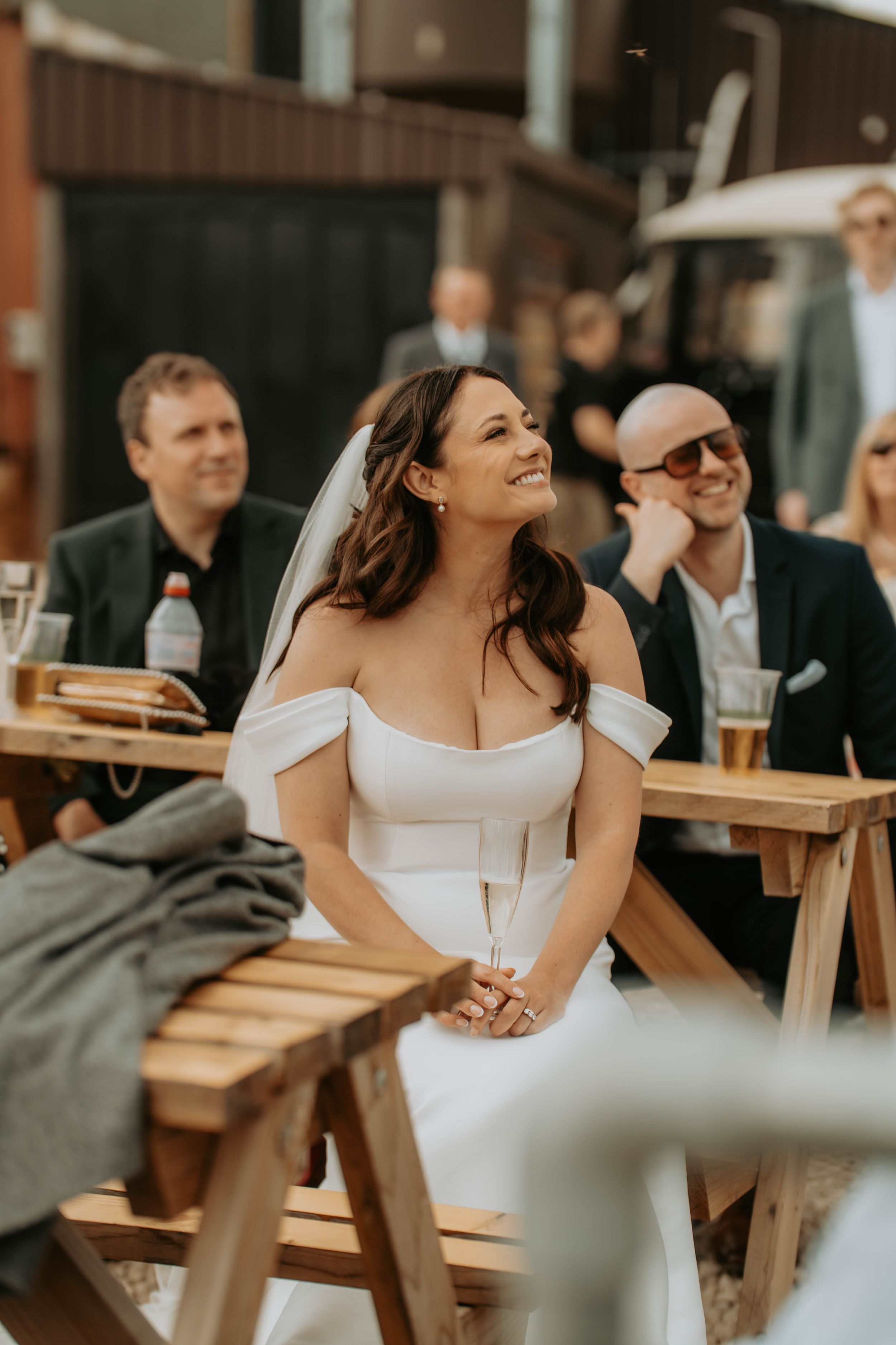 A smiling bride in an off-shoulder white gown and veil holds champagne at an outdoor wedding reception.