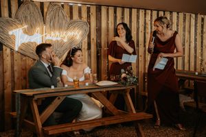 Bride and groom signing wedding documents at rustic wooden table with bridesmaids in burgundy dresses reading speeches nearby.