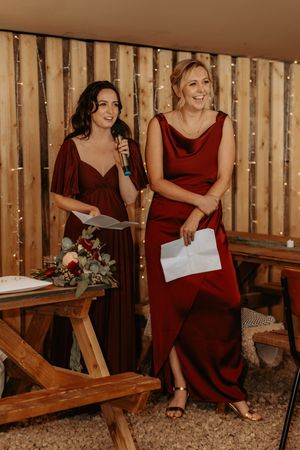 Two bridesmaids in burgundy dresses giving a speech at a rustic barn wedding with fairy lights.
