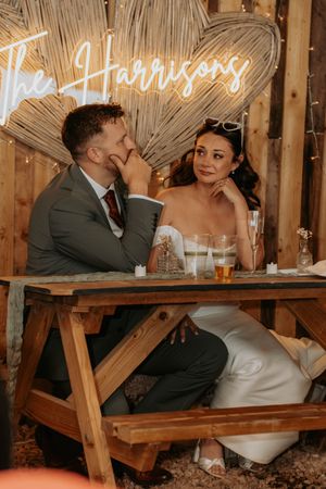 Bride and groom sit at rustic wooden table beneath glowing neon sign reading The Harrisons at their barn wedding reception.