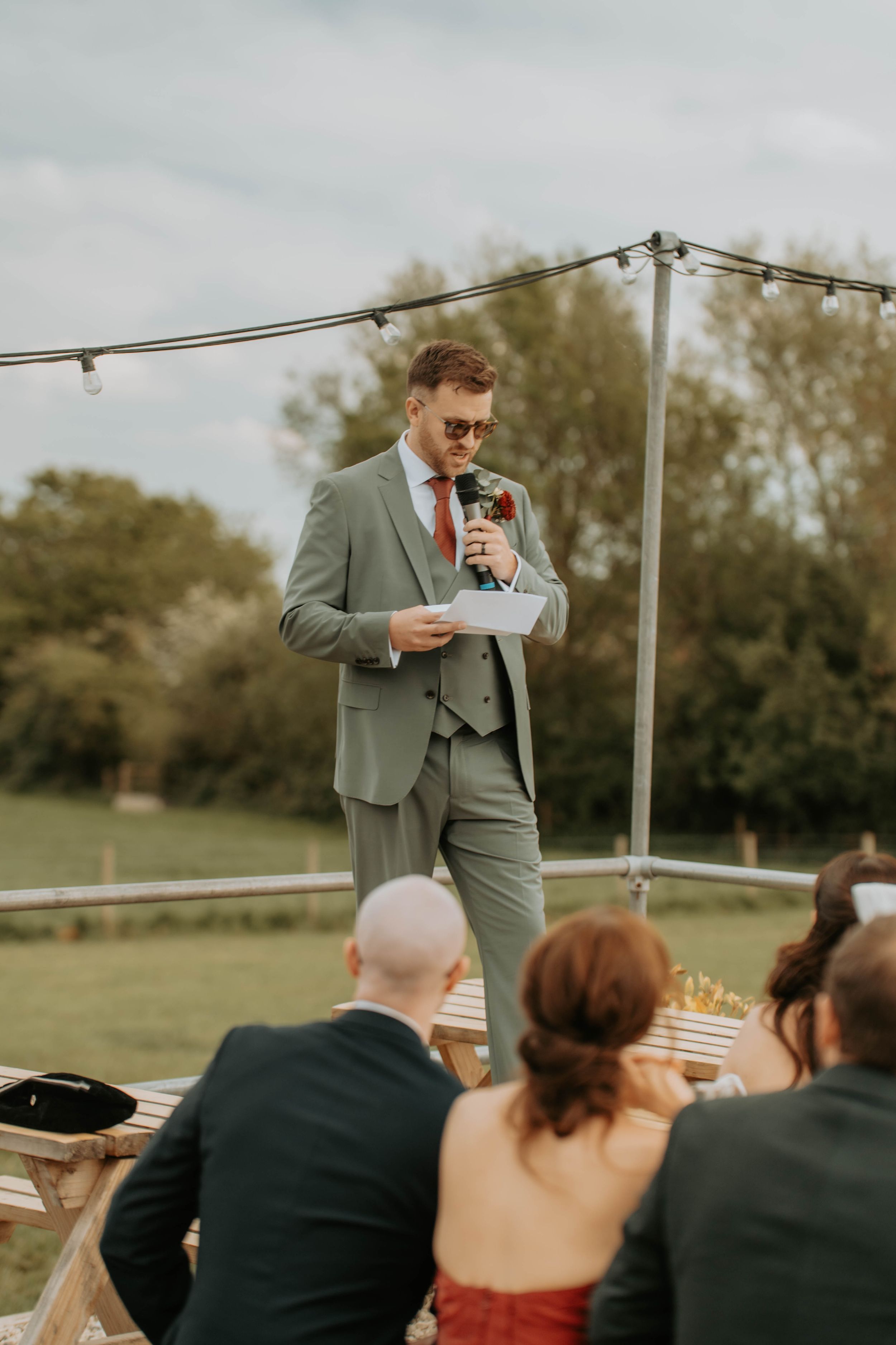 A groom in a gray suit delivers a speech outdoors at a wedding reception while guests listen attentively.