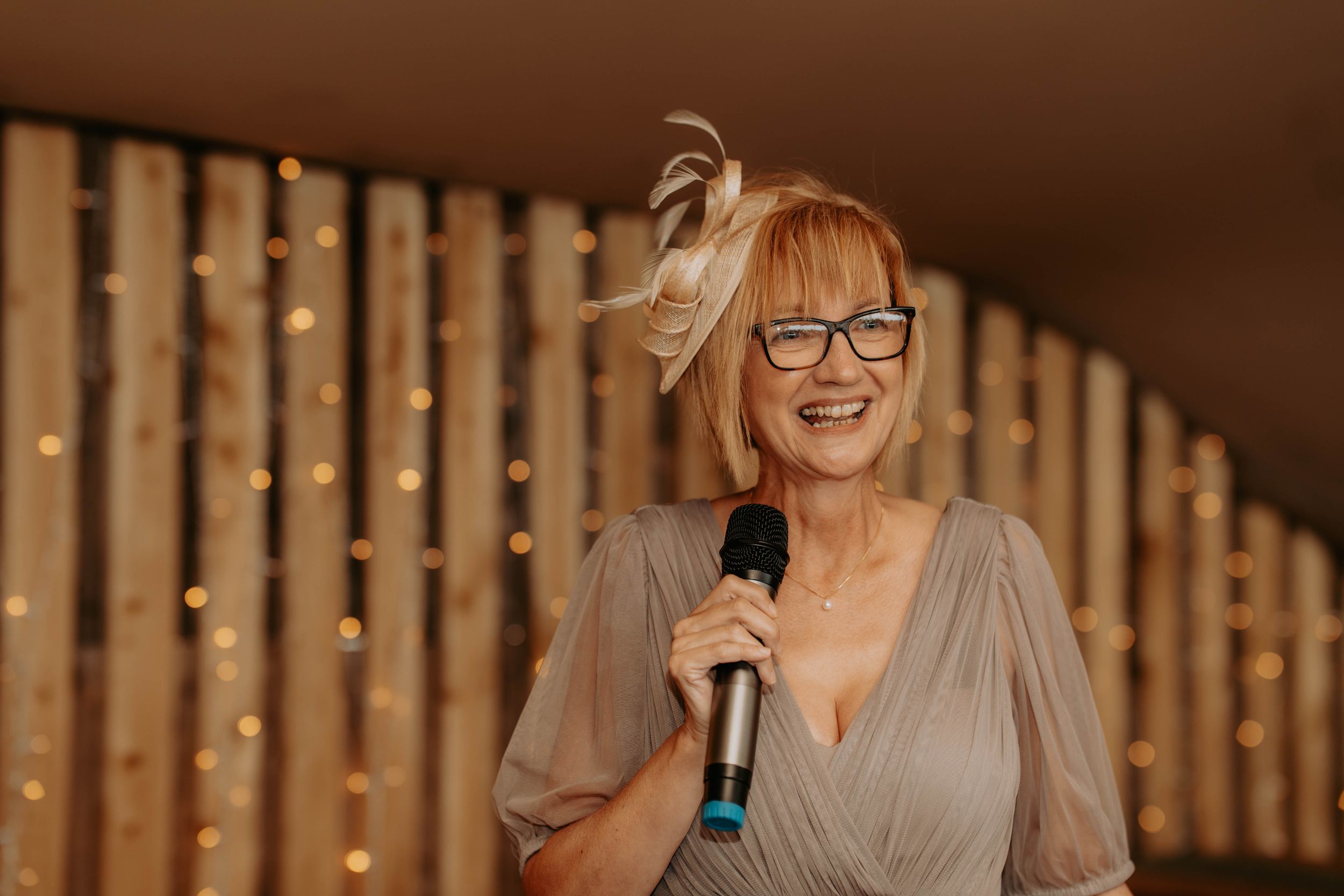 Woman with glasses and fascinator hat smiling while speaking into a microphone at a wedding reception.