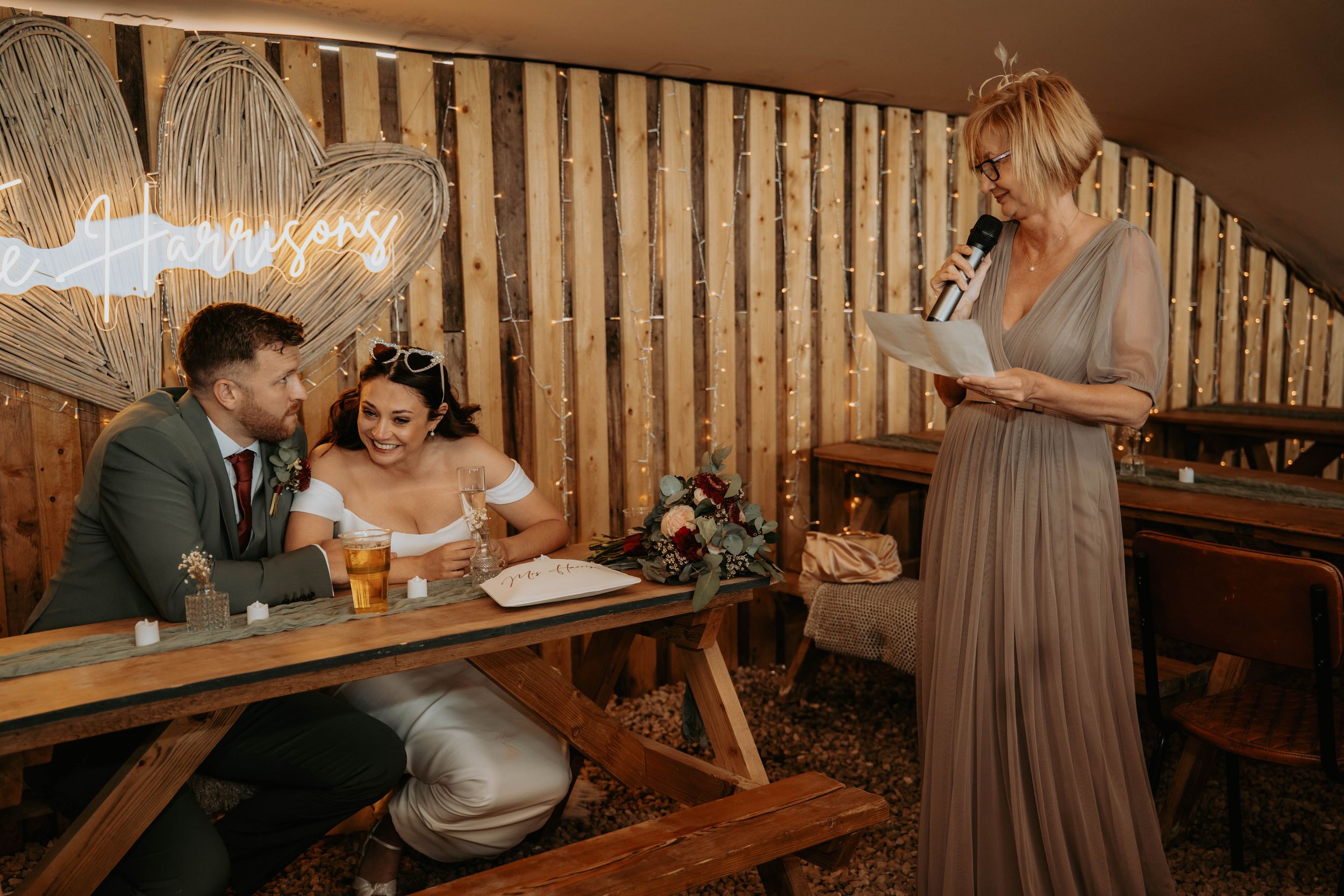 Bride and groom signing wedding register at rustic wooden table while bridesmaid reads speech in cozy venue.