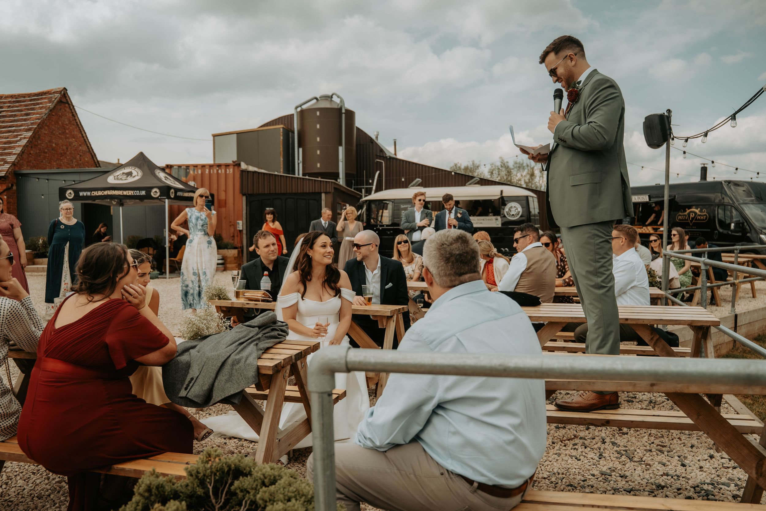 Outdoor wedding reception with guests seated at picnic tables while a man in a suit delivers a speech.