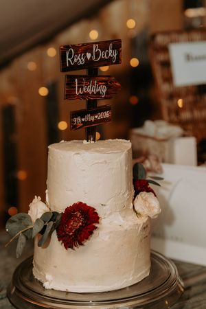 Two-tier white wedding cake with red and white flowers, rustic wooden topper reading Ross & Becky Wedding 9th May 2026.