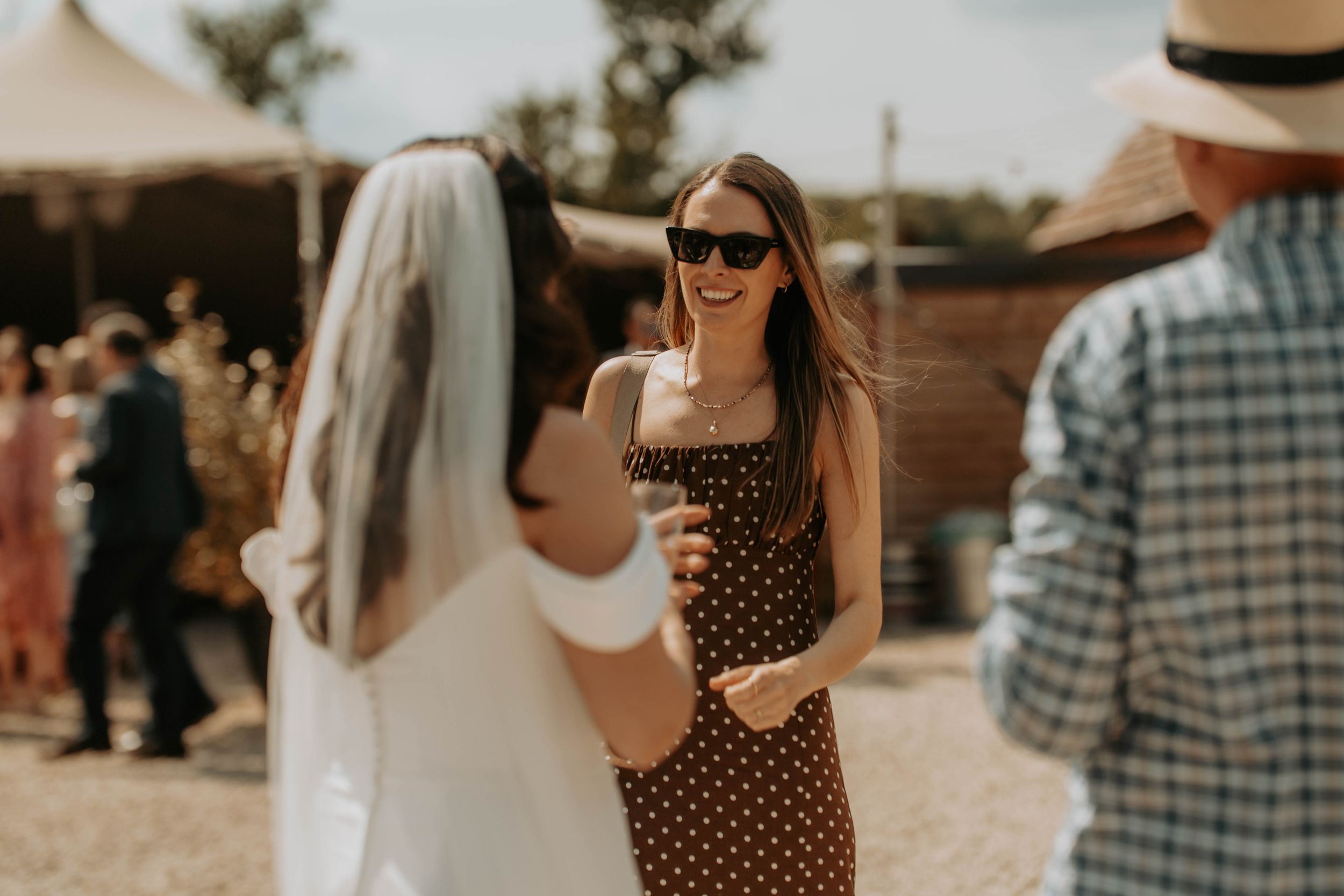 Woman in polka dot dress and sunglasses smiling with bride at outdoor wedding celebration.