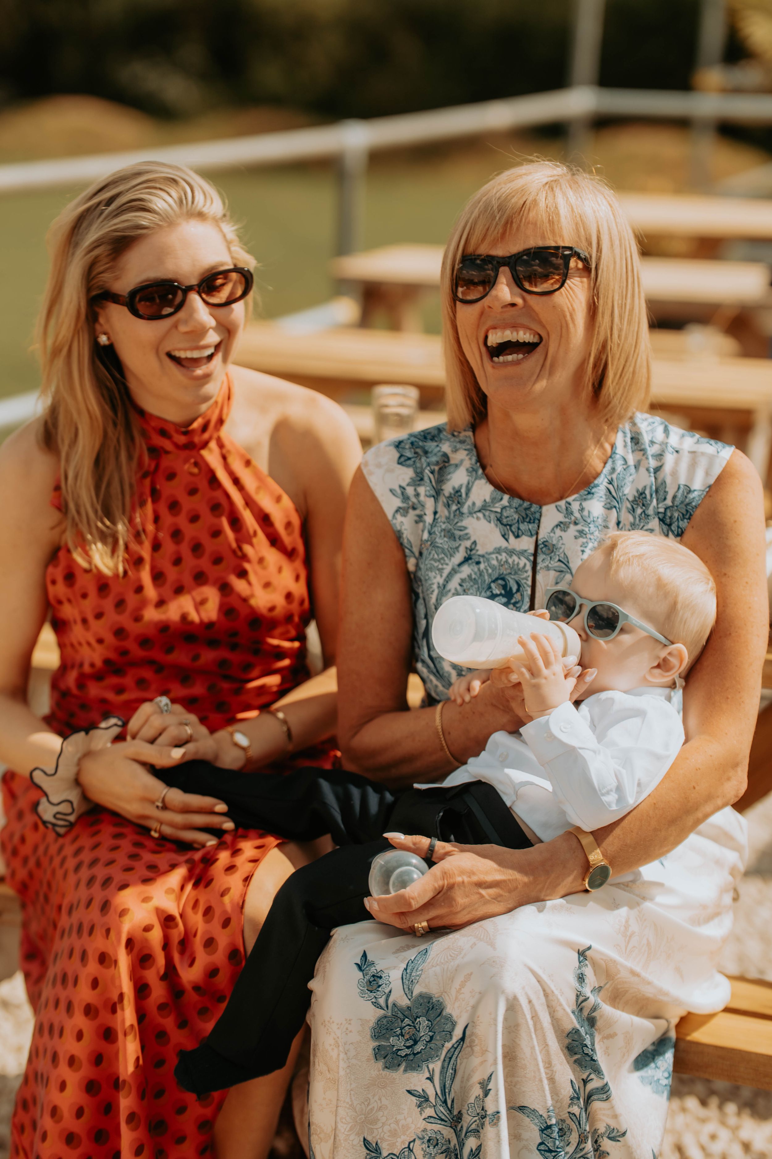 Two women in sunglasses laughing outdoors, one holding a baby in white being bottle-fed at a sunny gathering.