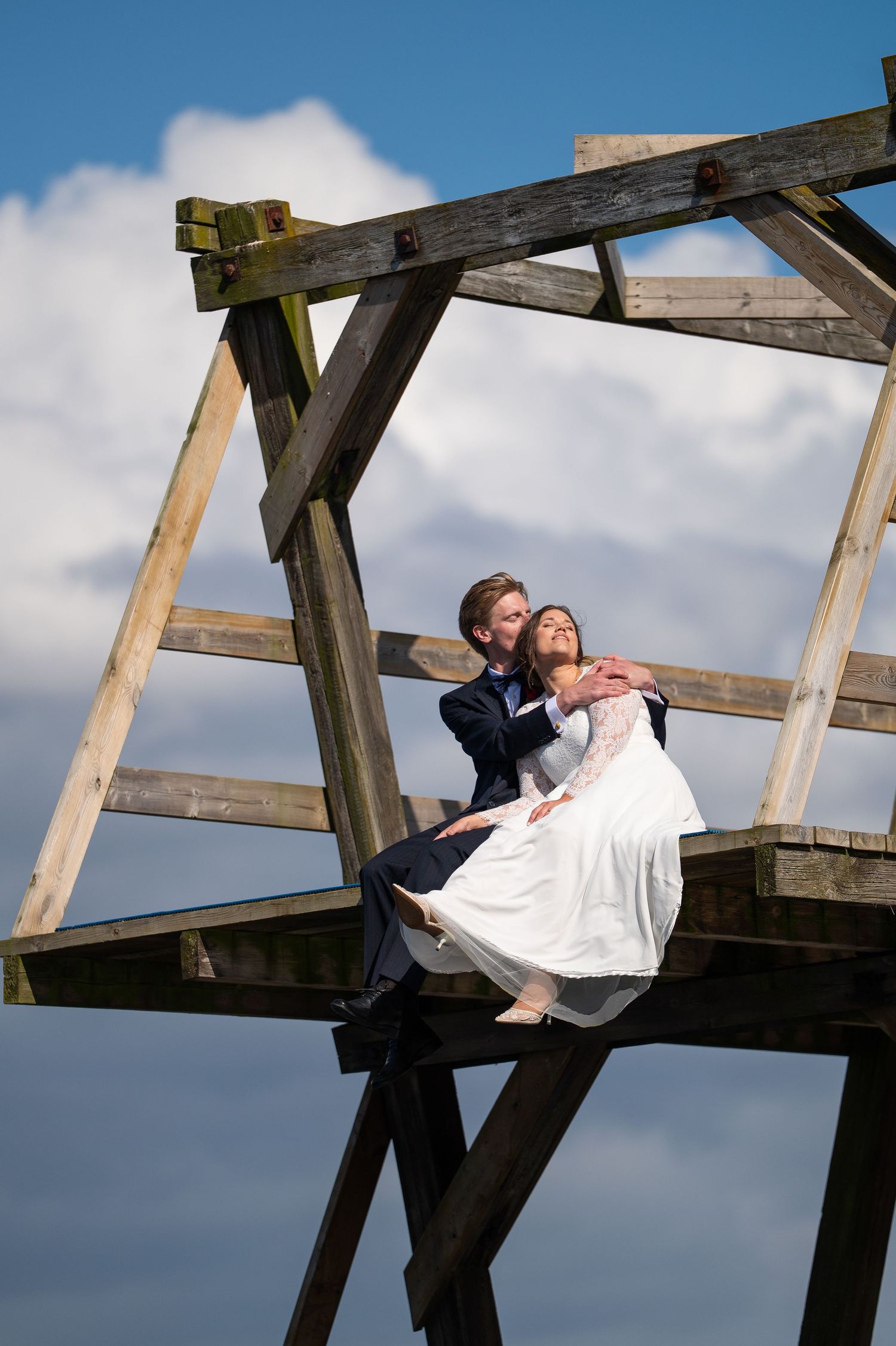 Bride and groom embrace romantically atop a rustic wooden structure against a blue sky on their wedding day.