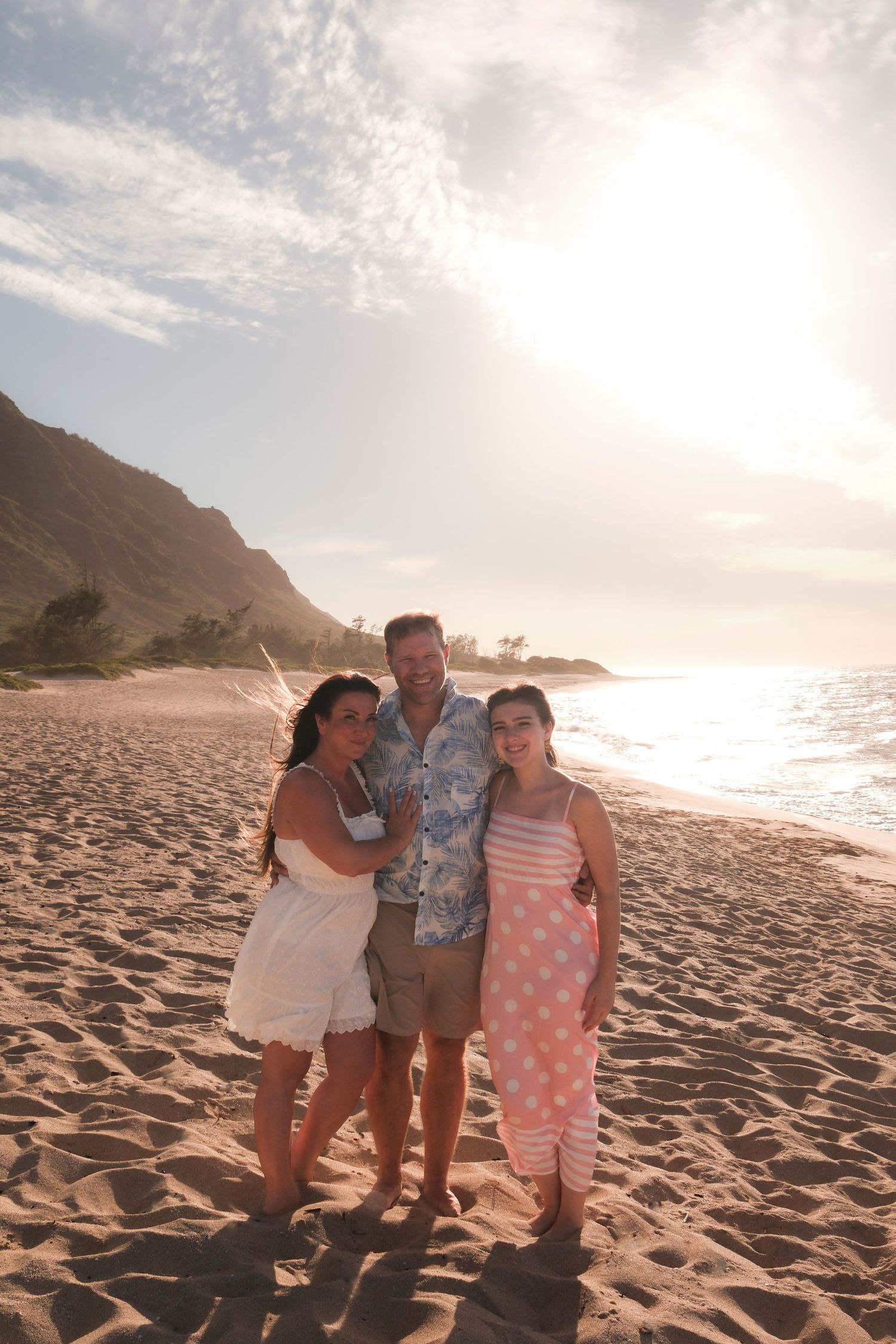 Family of three embracing on a North Shore Oahu beach surrounded by mountains and soft evening light