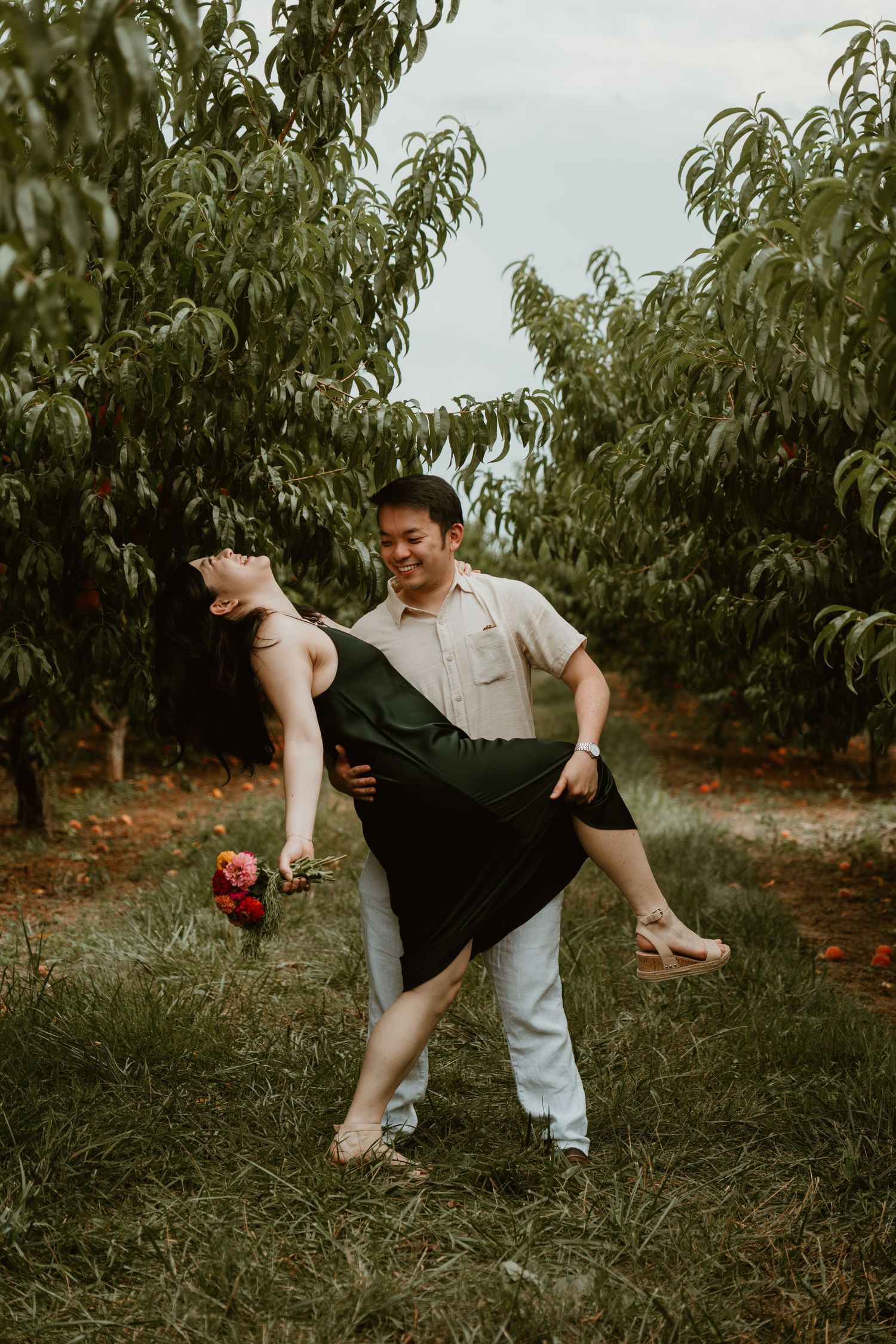 Couple dancing joyfully in a peach orchard, man lifting woman in black dress holding pink flowers.