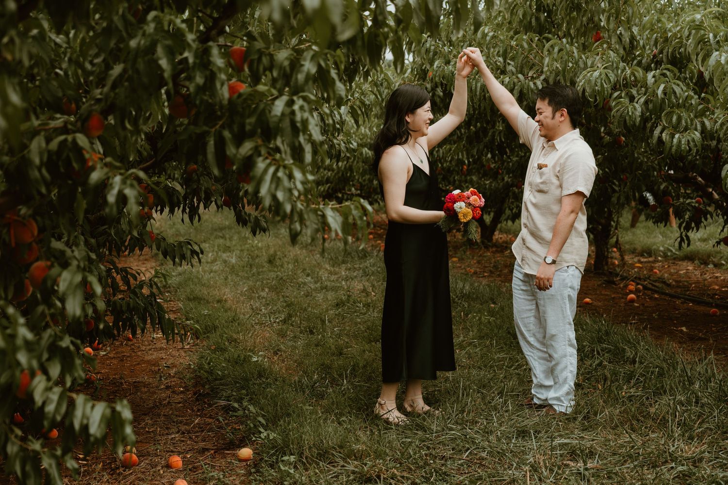 Couple dancing in a peach orchard, woman in black dress holding colorful bouquet, man in white shirt, surrounded by fruit trees.