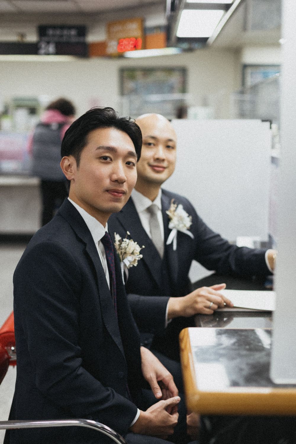 Two grooms in dark suits with boutonnieres sitting at a counter, smiling during their wedding registration ceremony.
