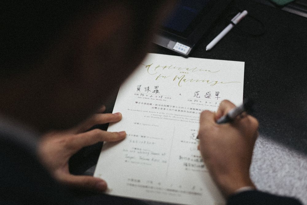 Person signing a marriage application form with Chinese characters and elegant calligraphy title at a desk.