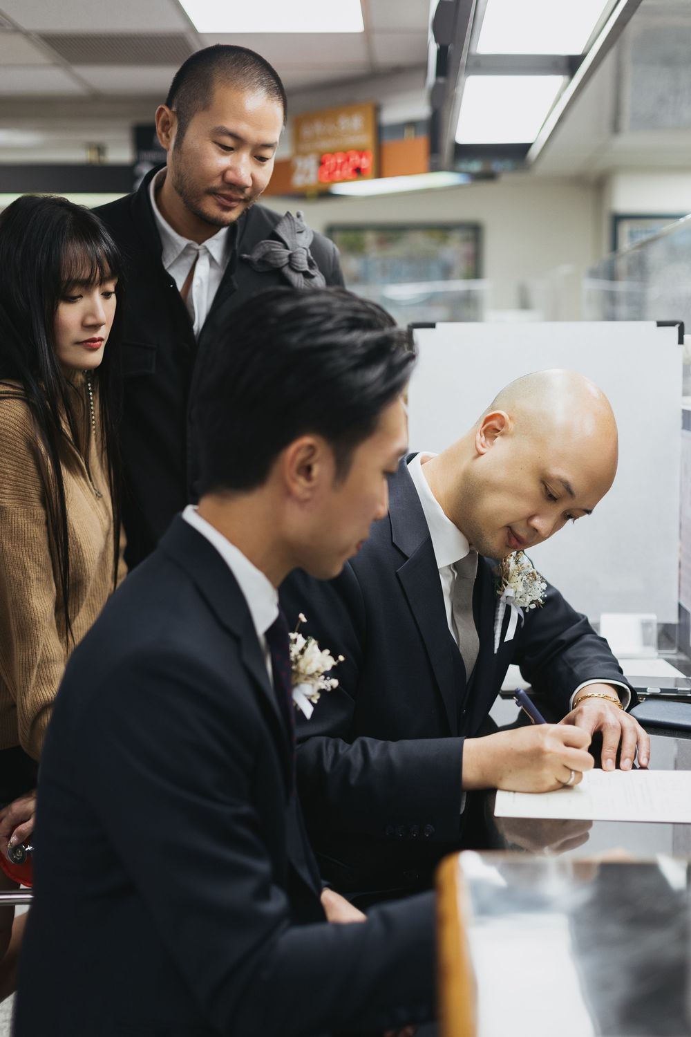 Two grooms sign marriage documents at a counter while witnesses look on during a civil wedding ceremony.