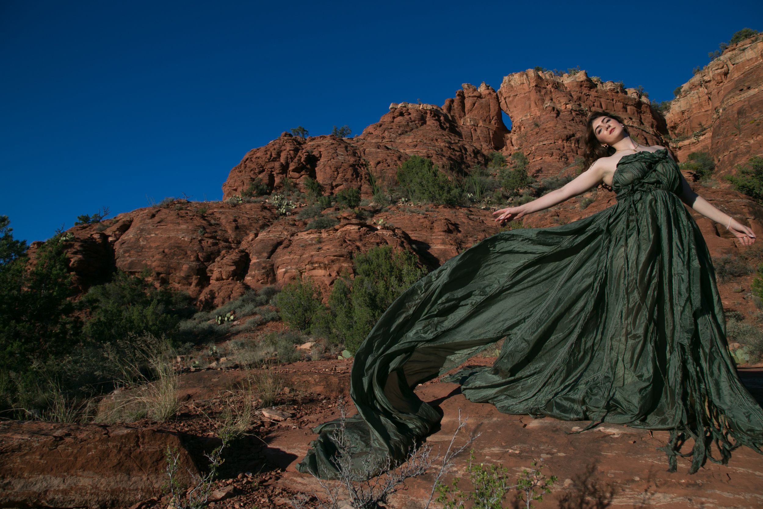 Woman in flowing green gown poses dramatically against red rock canyon landscape under vivid blue sky.