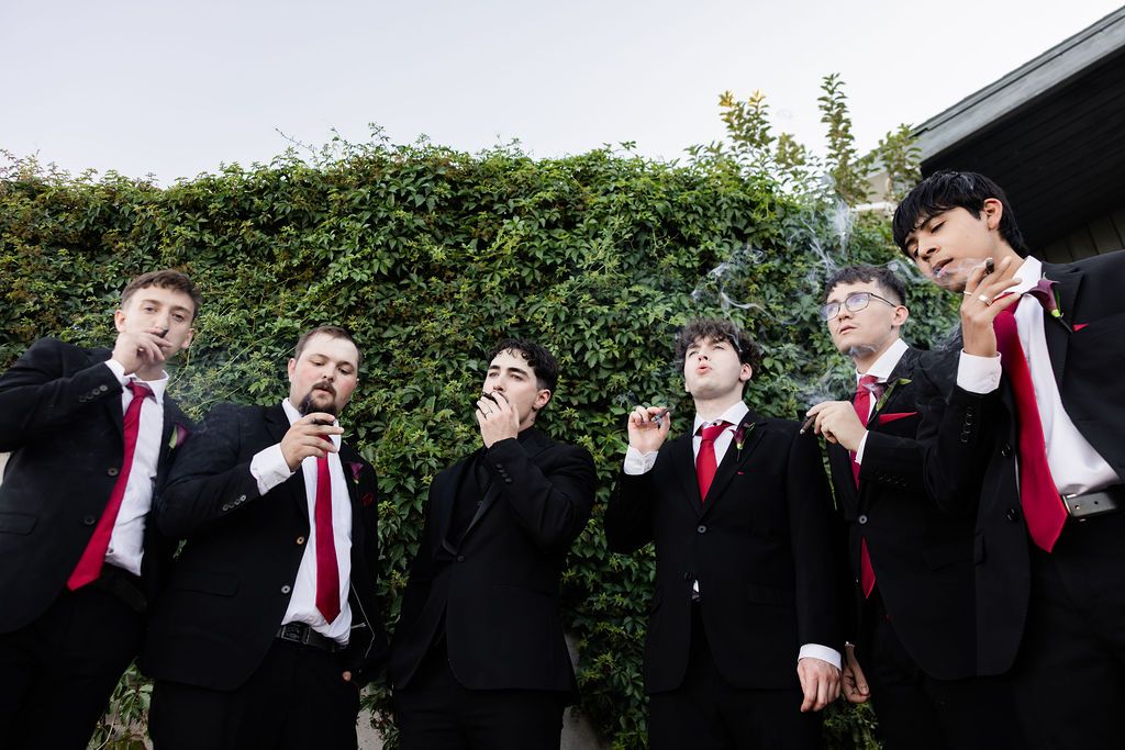 Six young men in black suits with red ties smoking cigars outdoors near green hedges.