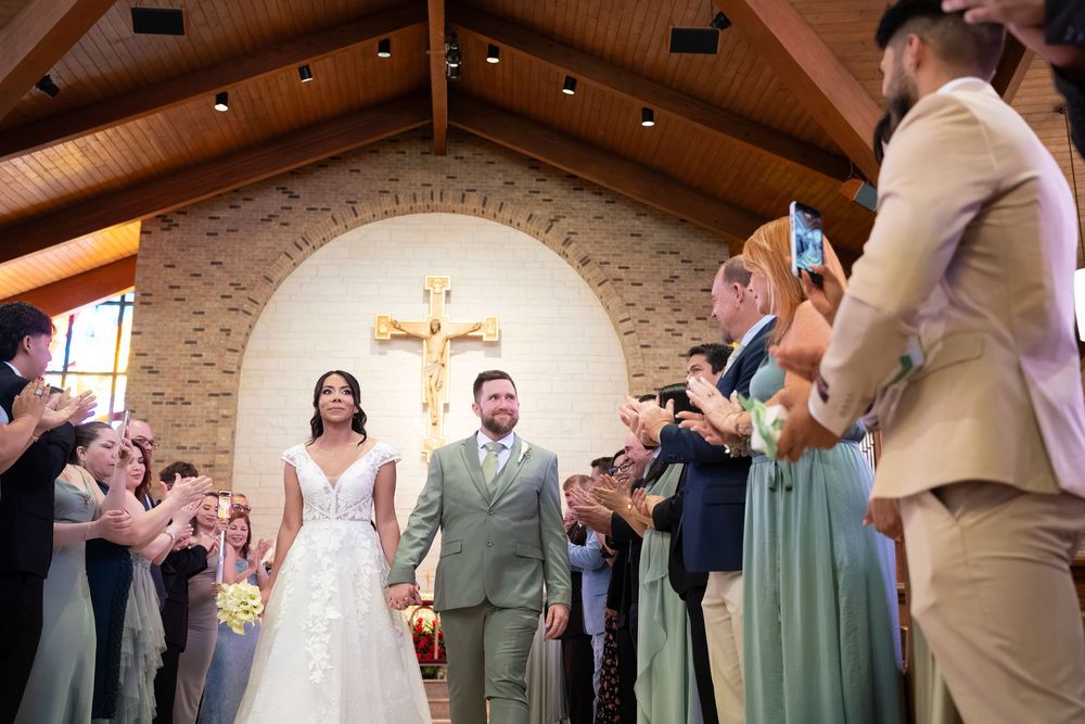 Newlywed couple walks down church aisle after wedding ceremony, guests applauding, cross visible at altar.