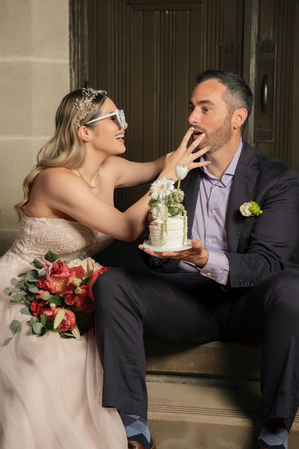 Bride and groom sitting on steps, sharing wedding cake, bride wearing tiara and sunglasses, holding red bouquet.