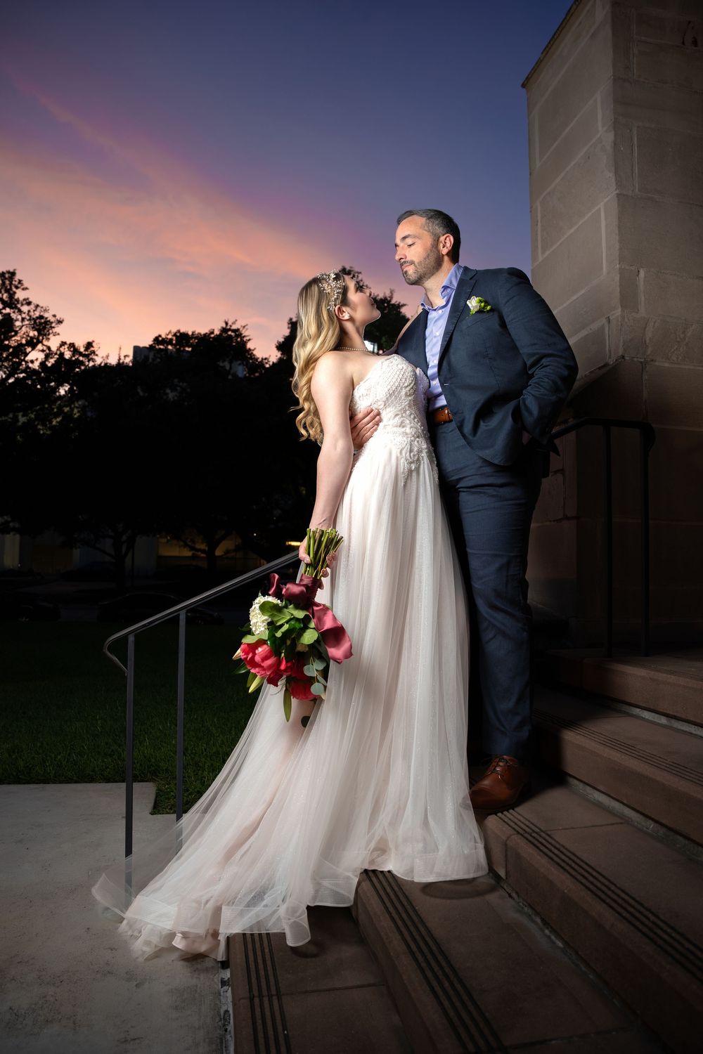 Bride and groom share a romantic kiss on steps at sunset, she holds red bouquet in flowing white gown, he wears navy suit.