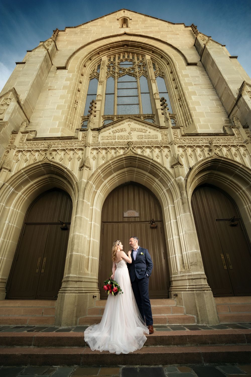 Bride and groom embrace before grand Gothic stone church entrance on wedding day.