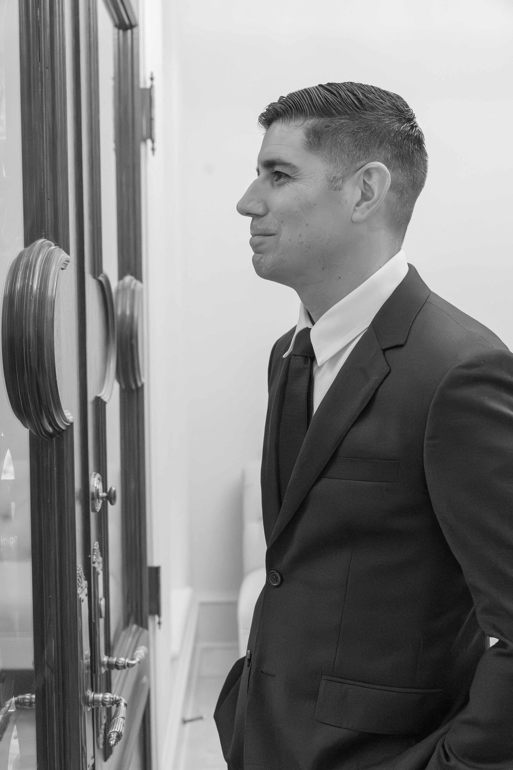Black and white photo of a man in a dark suit and tie standing in profile view near an ornate door.