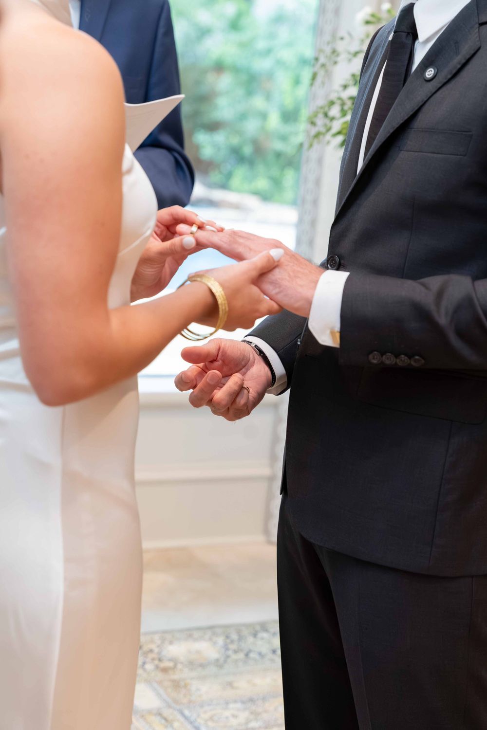 Bride placing a gold wedding ring on groom's finger during a wedding ceremony, both dressed in formal attire.
