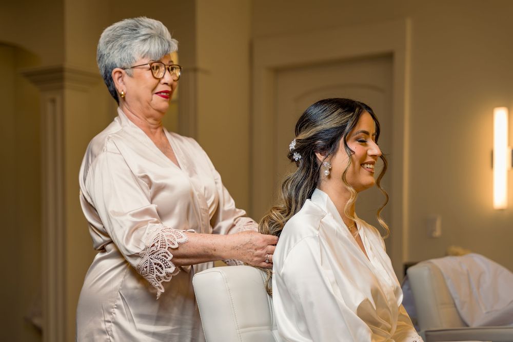 Older woman in satin robe helping smiling bride get dressed during wedding preparations in elegant room.