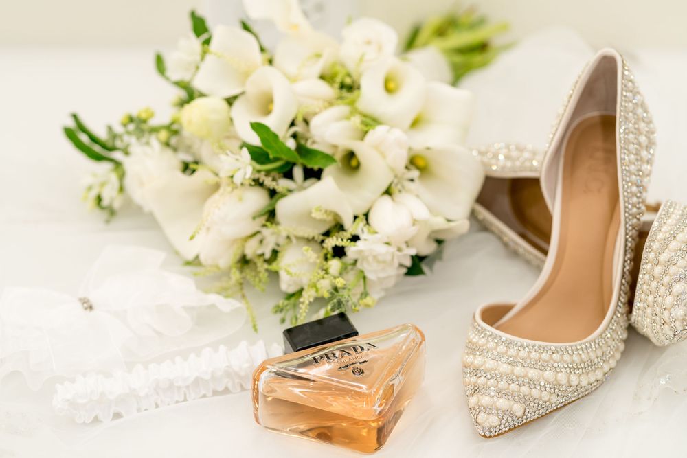 Pearl-embellished bridal heels beside a white floral bouquet and Prada perfume bottle on a white surface.