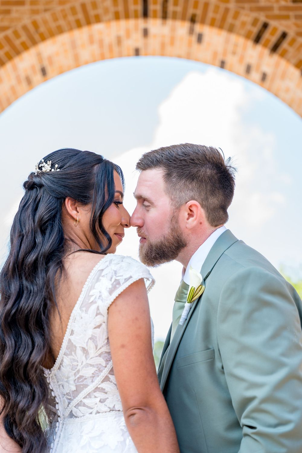Bride and groom sharing a romantic almost-kiss under a brick arch, bride in lace gown, groom in sage suit.