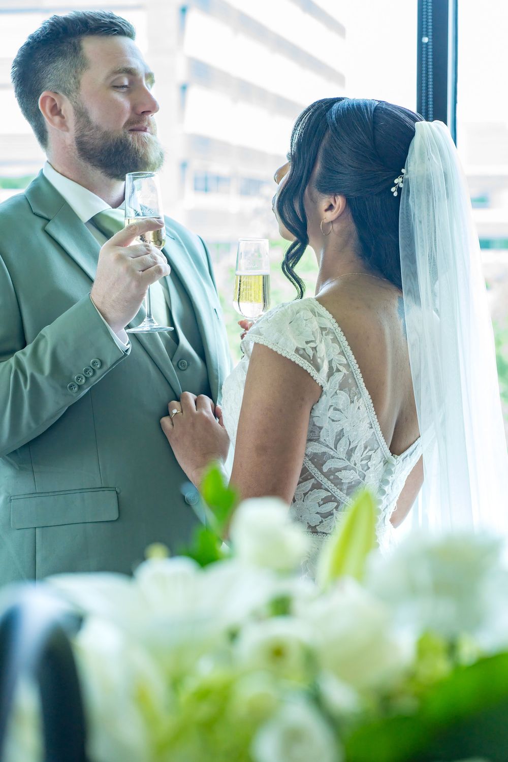 Bride and groom toasting with champagne glasses in wedding attire, white floral arrangement in foreground.