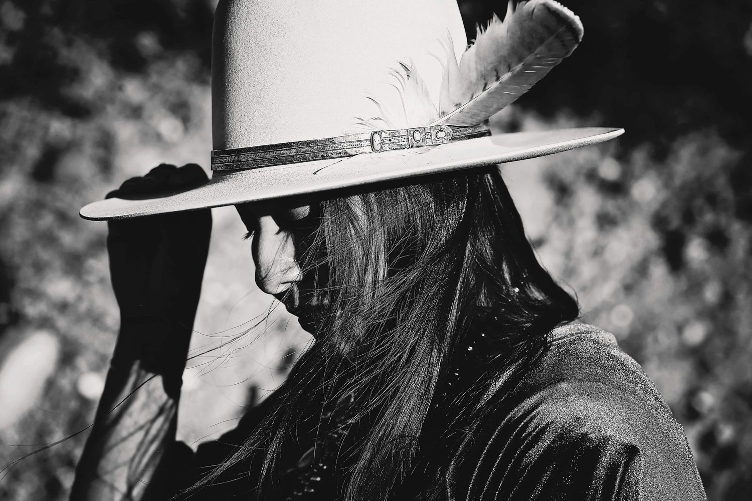 Black and white portrait of a person with long dark hair in profile, wearing a wide-brim hat with a feather and a beaded necklace.