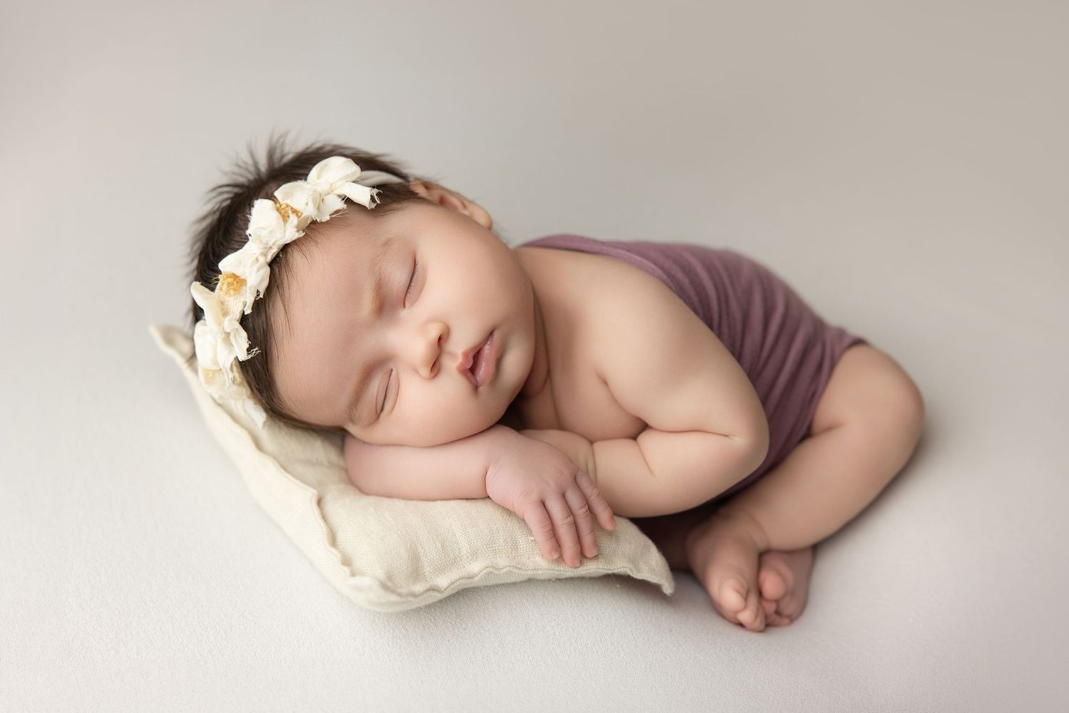 Sleeping newborn baby girl in purple wrap with floral headband resting on cream pillow.