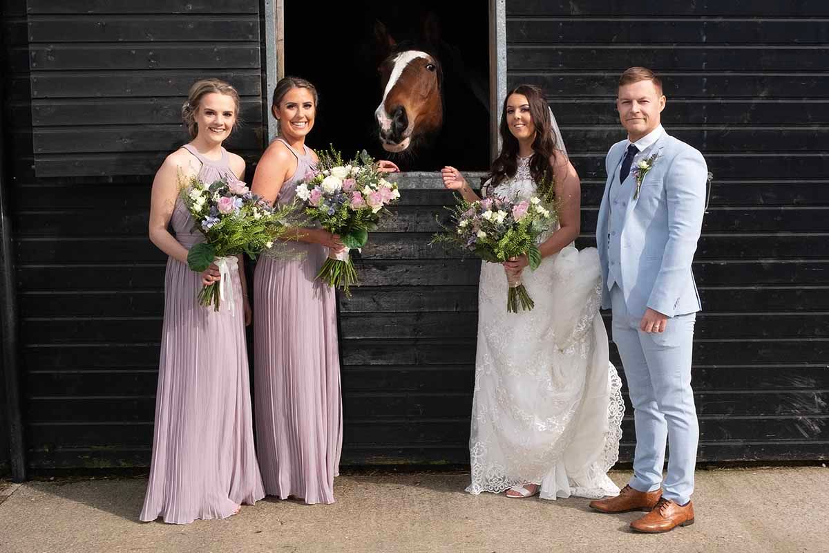 Bride and groom pose with two bridesmaids holding floral bouquets beside the Cart Shed at New House Farm wedding venue Lorton Cockermouth