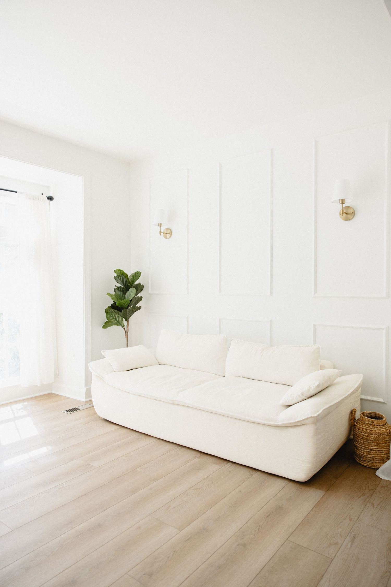 Minimalist living room with white sofa, fiddle leaf fig plant, gold wall sconces, and light wood floors.