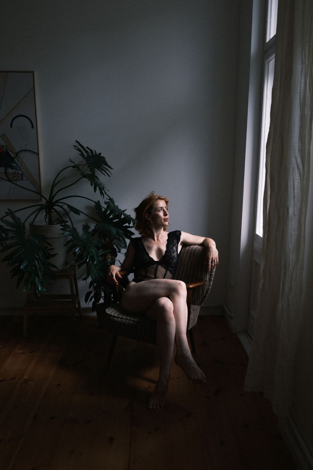 Woman in black lingerie sitting in wicker chair by window with natural light and indoor plants in moody room.